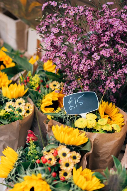 A lively display of fresh floral arrangements featuring bright yellow sunflowers, small white daisies with yellow centers, and red accent flowers wrapped in brown craft paper, set against a backdrop o