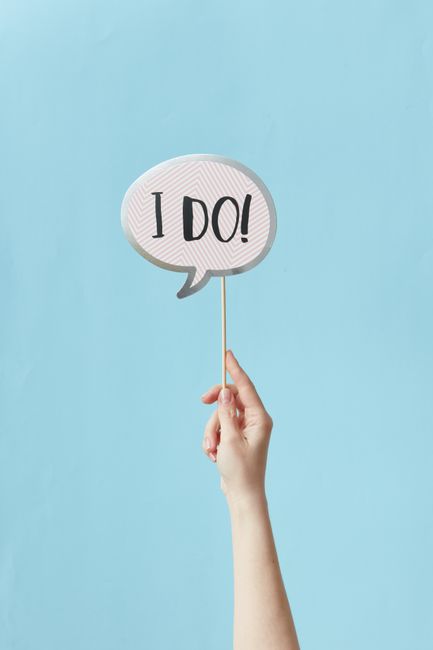 A person's hand holding a small, white speech bubble sign with the text 'I DO!' written in bold black letters. The sign is attached to a thin wooden stick and held against a plain light blue backgroun