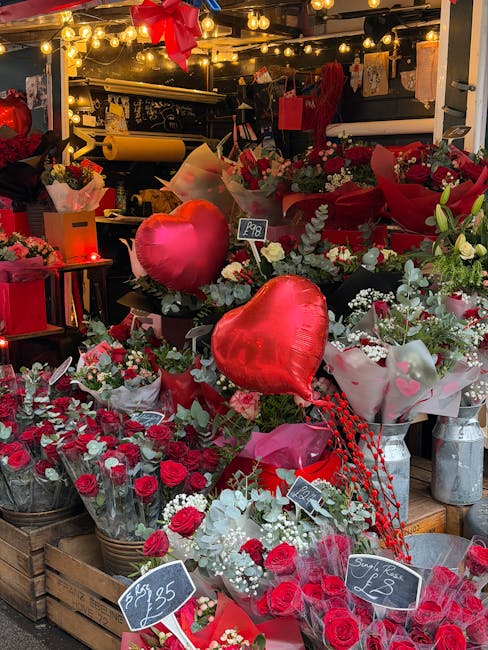 A vibrant display of fresh flowers and floral gifts at a florist shop, featuring red roses, white baby's breath, and greenery arranged in bouquets and wrapped in pink, white, and red paper. The arrang