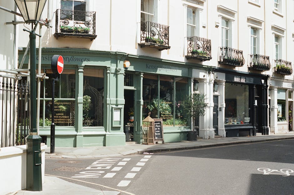 A view of a florist shop with a pastel green facade on a street in Churchill Gardens, Pimlico, featuring large glass windows displaying potted plants and floral arrangements. The shop has black and wh