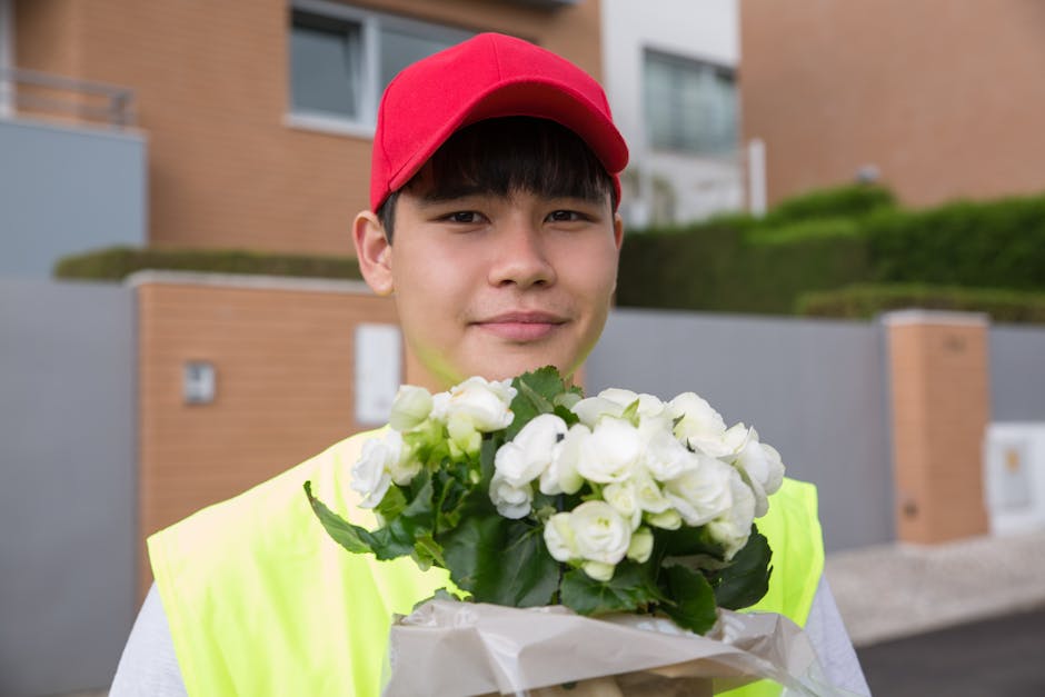 A young male flower delivery courier wearing a red cap and neon yellow vest holding a bouquet of white roses with green leaves. The bouquet is wrapped in light-colored paper. The courier stands outdoo