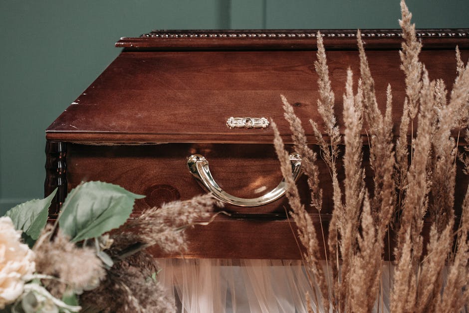 A polished wooden coffin with silver handle details, surrounded by tall, beige pampas grass and soft pale green leaves, is set against a dark teal wall. The arrangement suggests a floral tribute or me