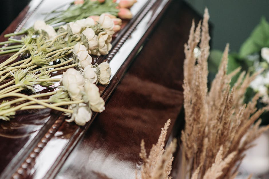 A floral arrangement consisting of white and soft peach roses with green foliage, placed on top of a polished dark wooden funeral casket. The flowers are arranged in a loose, natural style, with some 