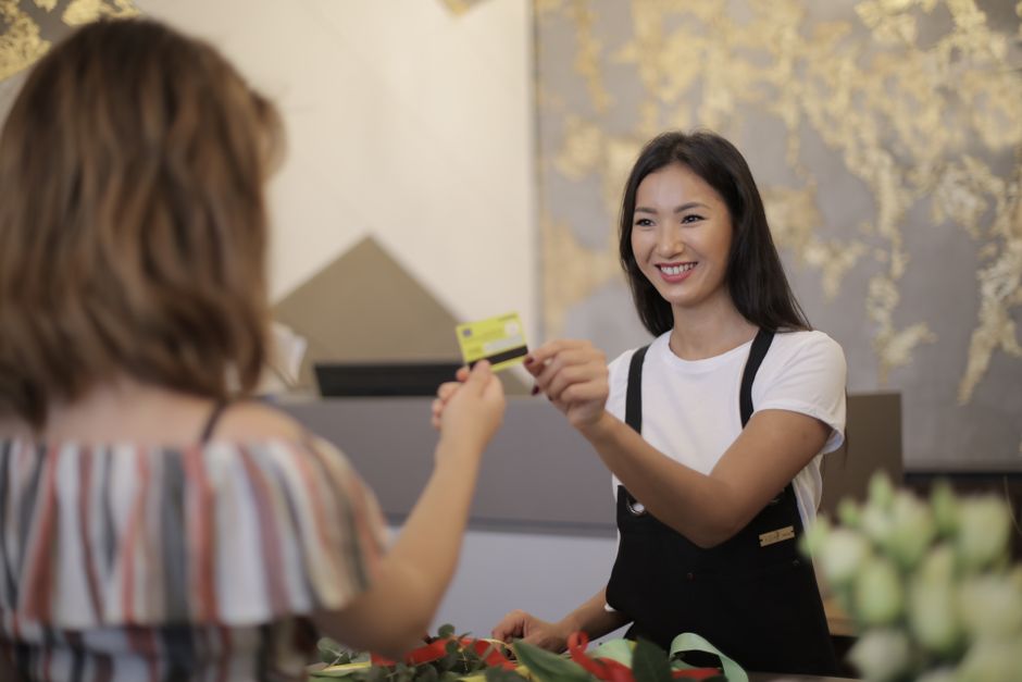 A young female florist with long dark hair, wearing a white t-shirt and black apron, is smiling as she hands a customer a floral gift card inside a florist shop. The customer, with shoulder-length bro