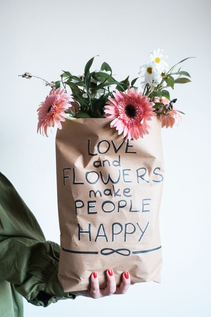 A bouquet of fresh flowers arranged in a brown paper bag with handwritten text that reads, 'LOVE and FLOWERS make PEOPLE HAPPY'. The bouquet includes large pink gerbera daisies with dark centers, smal