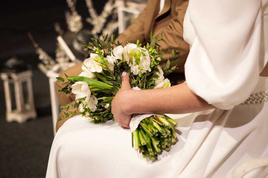 A close-up of a person holding a fresh bridal bouquet, featuring white lisianthus, with green foliage and long-stemmed flowers wrapped in white lace, resting on the person's lap. The background shows 