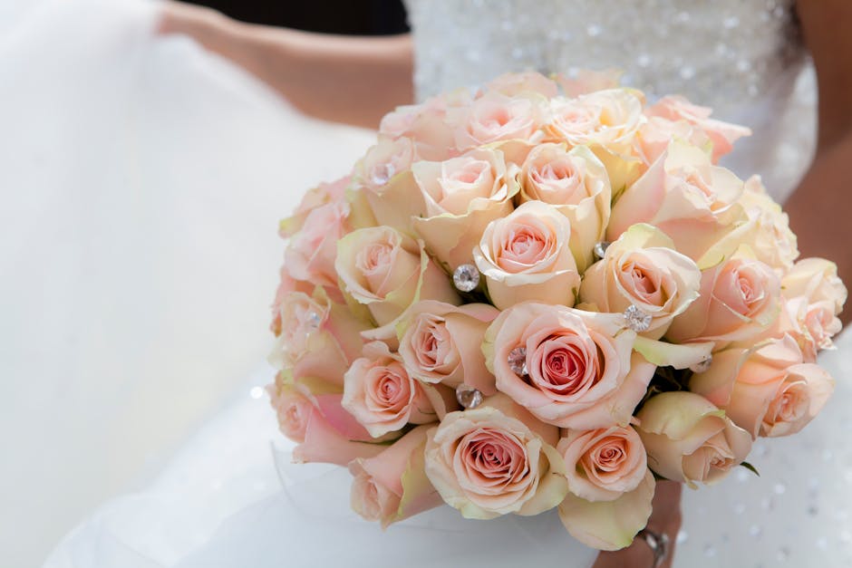 A close-up of a bridal bouquet composed of pale pink and cream roses with soft, velvety petals, arranged in a compact rounded shape. The bouquet includes accents of small, sparkling rhinestone embelli